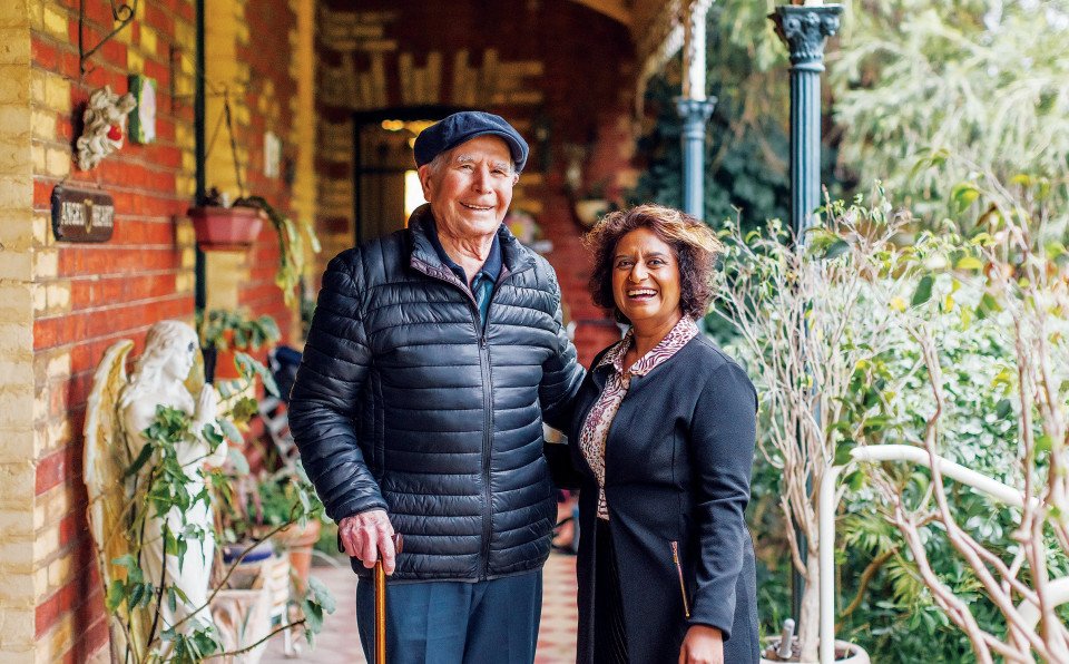 An old man and younger south asian woman stand on veranda of house smiling